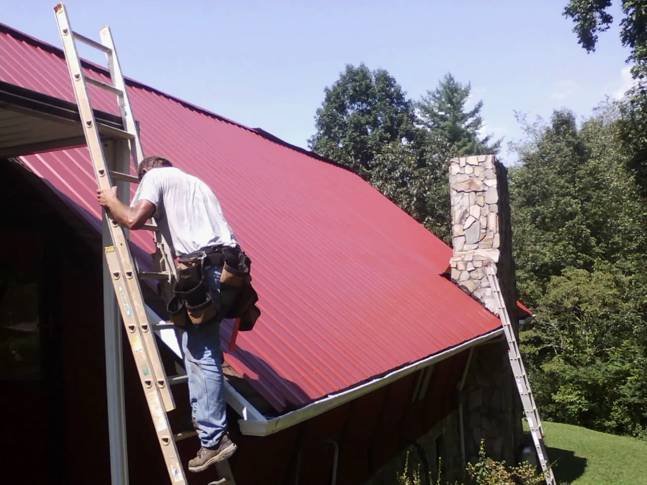 Roofer on a ladder beside a red metal roof and stone chimney — Soffit & Fascia Repair by Roofing Brothers in Bloomingdale